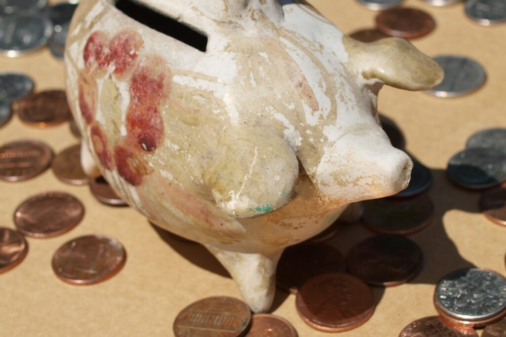 Cofrinho de cerâmica cercado de moedas sobre fundo marrom, representando guardar a primeira parcela do 13º salário