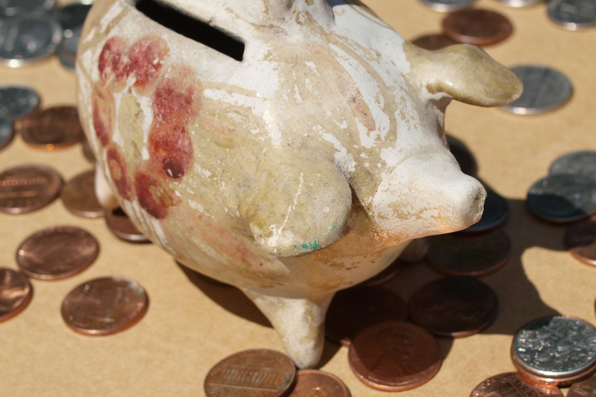 Cofrinho de cerâmica cercado de moedas sobre fundo marrom, representando guardar a primeira parcela do 13º salário