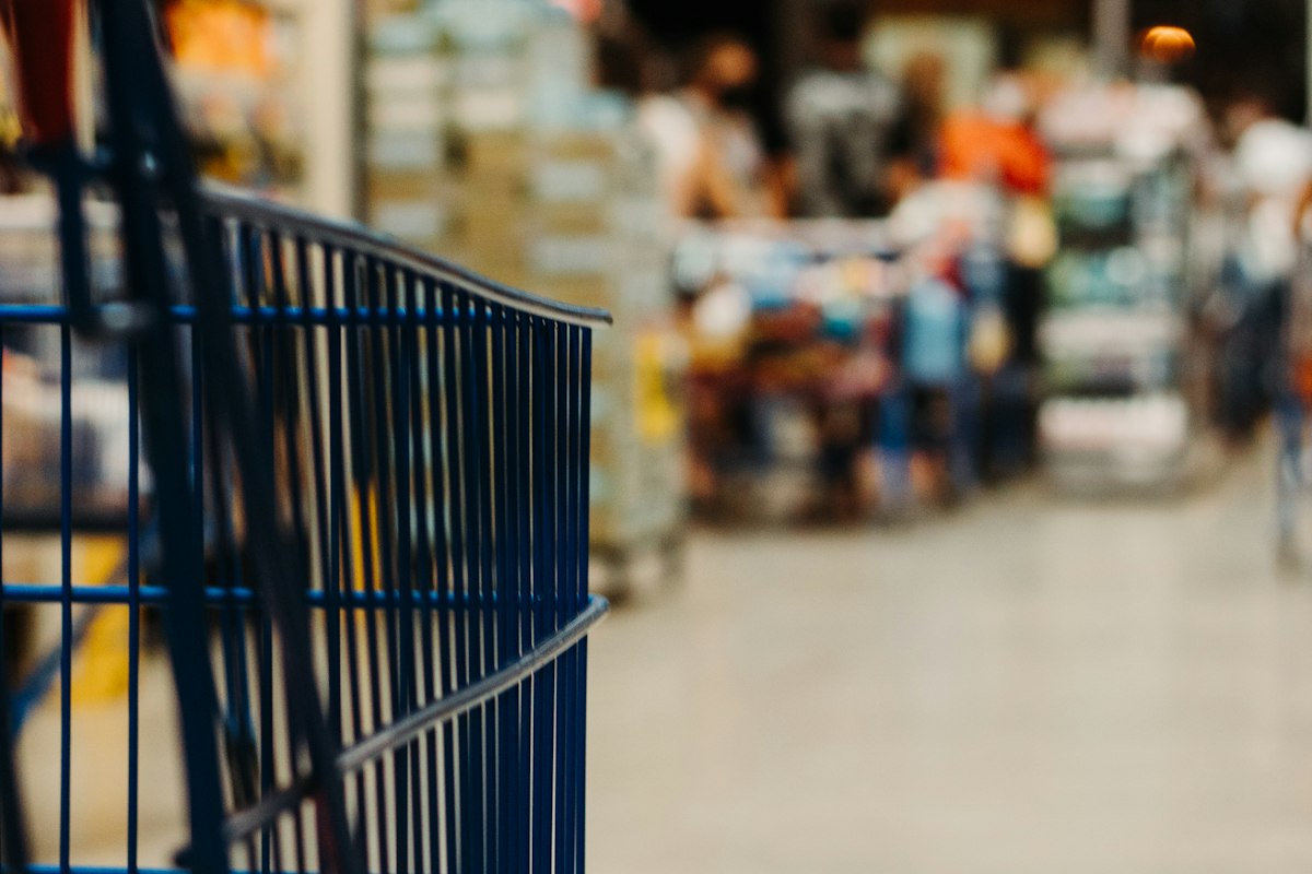 Carrinho de compras azul em primeiro plano num corredor de supermercado desfocado, representando a inflação no dia a dia