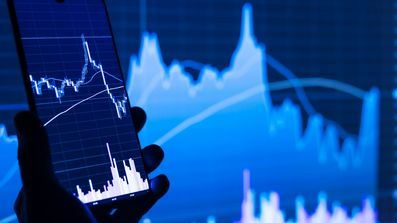 Pessoa segurando celular diante de um gráfico de mercado